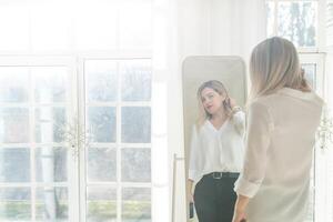 Self-confident Woman looking at her reflection into the mirror indoors. Beautiful interior design photo