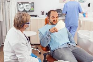 enfermo paciente inspeccionando dientes después dental cirugía utilizando espejo mientras sentado en estomatológico silla en hospital clínica oficina. mayor estomatólogo mujer que se discute acerca de cuidado de la salud tratamiento foto