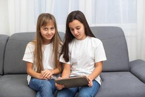 Cute little girl and older sister playing together smiling and having bonding time using a laptop on couch at home. Happy family Siblings relationship and digital technology lifestyle concept. photo