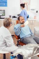 Senior stomatologist woman finishing dental surgery for man patient sitting on stomatology chair looking at tooth. Doctor explaining toothache treatment during stomatological consultation photo