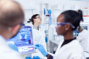 hembra científico sostiene matraz con cura para vacuna durante global pandemia. multiétnico equipo de médico investigadores trabajando juntos en estéril laboratorio vistiendo proteccion lentes y guantes. foto