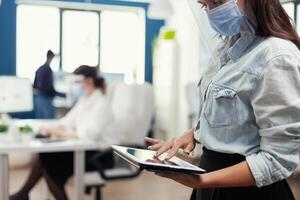 Employee from marketing department checking company statistics using tablet pc. Multiethnic coworkers respecting social distance in business company during coronavirus pandemic. photo