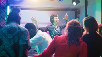 Young DJ mixing funky music at party, feeling cheerful with dance moves and electronic sounds at station on stage. Group of friends partying and dancing together at club, nightlife. photo