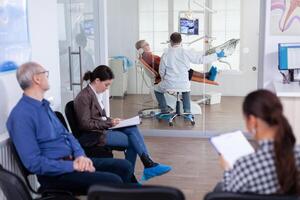 Crowded stomatology waiting area with people filling form for dental consultation. Stomatoloy specialist denstiry treating senior woman cavity. Receptionist working on computer. photo