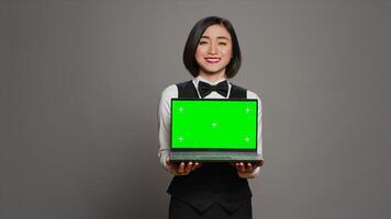 Asian hotel concierge holding pc with greenscreen on camera, presenting isolated mockup template on screen. Receptionist showing laptop with blank chromakey layout on display. Camera A. photo