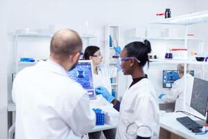 Scientist holding glass flask with blue genetic material in busy laboratory. Multiethnic team of medical researchers working together in sterile lab wearing protection glasses and gloves. photo