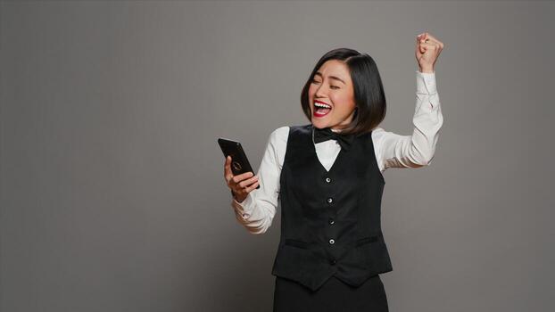 Front desk staff in uniform looks at online information on mobile phone, checking all website reservations over grey background. Asian receptionist using smartphone for bookings. Camera A. photo