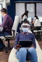 African manager taking notes on tablet pc wearing face mask against covid19 at workplace. Multiethnic business team working respecting social distance during global pandemic with coronavirus. photo