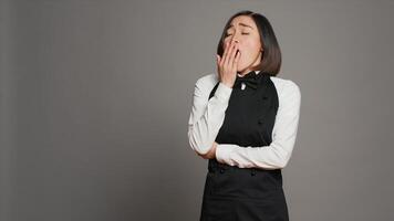 Exhausted waitress with apron yawning over grey background, feeling overworked and dealing with burnout after multiple restaurant events. Asian catering worker feeling tired in studio. Camera A. photo