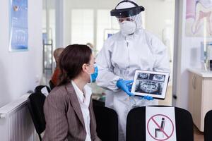 Stomatologist in protective suit pointing on digital x-ray of tooth explaining to patient treatment using tablet in covid-19 pandemic. Medical team wearing face shield, coverall, mask and gloves. photo