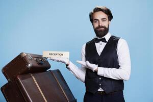 Bellboy holds reception sign on camera, indicating direction for guests arriving at hotel. Professional employee working in hospitality industry uses front desk logo, tourism. photo