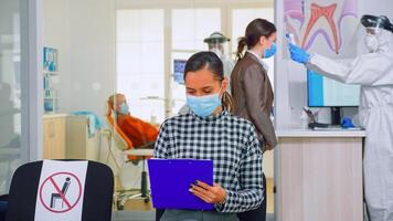 Woman with face protection mask writing on registration form in stomatomoly clinic, sitting in reception respecting social distance. Dentist working wearing overall in dental clinic with new normal photo