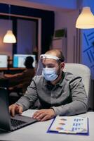 Manager and colleagues doing overtime with visor against covid-19. Stressed man in corporation working hard to finish a project wearing face mask as safety precaution due to coronavirus pandemic. photo