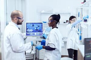 African assitant discussing wtih scientist about virus experiment in chemistry laboratory. Multiethnic team of medical researchers working together in sterile lab wearing protection glasses and gloves. photo