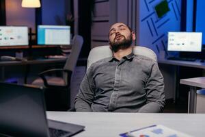 Overworked exhausted man sleeps on chair in empty office. Workaholic employee falling asleep because of while working late at night alone in the office for important company project. photo