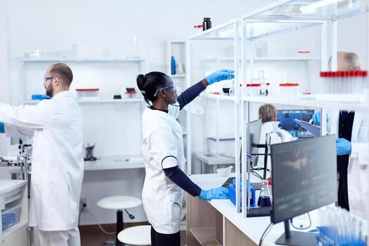 African in biotechnology laboratory reaching for glass flask from shelf. Multiethnic team of researchers working in microbiology lab testing solution for medical purpose. photo
