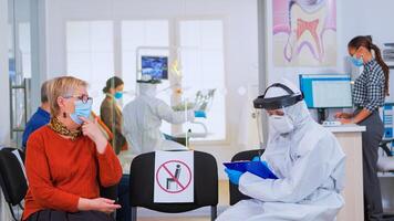 Dentist with face shield talking with elderly woman with toothache before examination during pandemic covid-19 sitting on chairs in waiting area keeping distance. Concept of new normal dentist visit photo