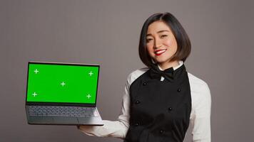 Restaurant employee pointing at greenscreen layout on pc, smiling at camera and showing isolated mockup screen. Asian waitress looking at blank copyspace display on computer. Camera B. photo