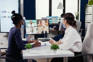 African employee and manager discussing on video conference with businesspeople wearing face mask for covid. New normal business office. Multiethnic team working respecting social distance during global pandemic with coronavirus. photo