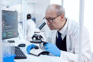 Senior man carrying out scientific investigation looking throug microscope. Chemist researcher in sterile lab doing experiments for medical industry using modern technology. photo