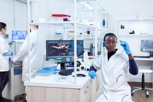 African biochemistry looking at sample on glass slide sitting at her workplace. Black healthcare scientist in biochemistry laboratory wearing sterile equipment. photo