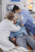 Specialist dentist team with face mask examining patient mouth during medical surgery in stomatological office room. Senior woman doctor using professional dentistry tools for cavity procedure photo