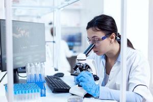 Microbiologist in research laboratory doing scientific experiments looking through microscop. Chemist wearing lab coat using modern technology during scientific experiment in sterile environment. photo