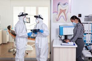 Stomatology team dressed up in ppe suit during global pandemic with coronavirus in dental reception holding patient x-ray, keeping social distancing. Receptionist with face mask as prevention. photo