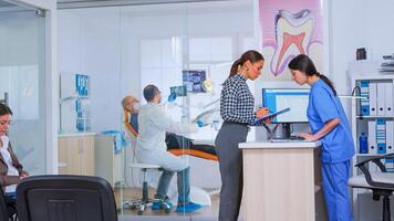 Professional dentist asking nurse for dental x-ray before examining patient while people waiting in reception area of modern stomatological clinic. Dental nurse typing on computer making appointments. photo