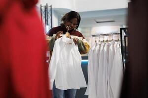 Clothing store buyer trying on shirt while exploring formal apparel for wardrobe. African american woman holding outfit on hanger, examining size, style and fit while shopping in boutique photo