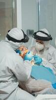 Stomatologist in protective suit performing dental examination taking care of teeth using sterilized tools. Medical team working wearing coverall, face shield, mask, gloves in stomatological office photo