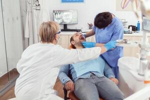 Sick man sitting on dental chair while medical nurse looking into mouth checking teeth health during stomatology consultation. Senior dentist woman preparing tools for oral surgery photo