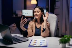 Businesswoman waving in the course of call with colleagues doing overtime. Woman working on finance during a video conference with coworkers at night hours in the office. photo