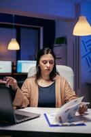 Focused entrepreneur looking at charts trying to finish deadline in empty office. Woman checking notes on clipboard while working on important project doing overtime. photo