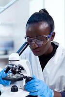 bioquímica técnico con africano etnia utilizando microscopio para farmacia industria con protector lentes. negro cuidado de la salud científico en bioquímica laboratorio vistiendo estéril equipo. foto