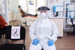 Close up of tired doctor in stomatological office looking on camera wearing overall and face shield sitting on chair in waiting room clinic. Concept of new normal dentist visit in coronavirus outbreak. photo