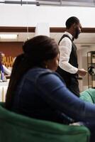 African American hotel staff and guest in lobby. Woman traveler waiting for check-in at reception area, friendly smiling bellboy helping guests with luggage. Hospitality and tourism Industry photo