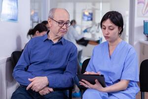 Nurse taking notes on tablet pc discussing with senior patient in dental office, explaining diagnosis. Assistant filling writing healthcare form. Dentistiry hospital. photo