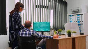 Entrepreneur pointing on desktop discussing with disabled colleague in new normal office in front of computer. Team of financial expert working with protection face masks respecting social distance. photo