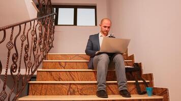 Overworked office executive sitting on stair typing on laptop looking on documents from clipboard while colleagues moving on staircase. Professional businesspeople working in modern financial building photo