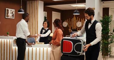 Guests checking out from resort, handing over room access card key while friendly bellboy helps them with their baggage. Happy relaxed tourists leaving hotel after nice stay photo