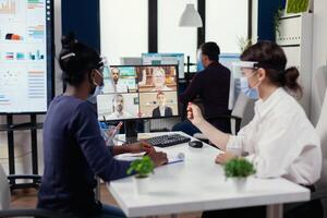 negro mujer y colega escuchando negocio personas en vídeo llamada vistiendo cara máscara durante COVID-19. nuevo normal negocio oficina. multiétnico equipo trabajando respecto a social distancia durante global pandemia con coronavirus. foto