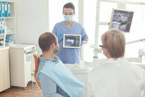 Stomatologist nurse pointing on digital screen explaining tooth x-ray to sick man sitting on dental chair in hospital stomatological office. Dentist doctor discussing toothache treatment photo