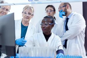 Group of scientists in medicine lab looking at computer display analysing virus. Black healthcare researcher in biochemistry laboratory wearing sterile equipment. photo