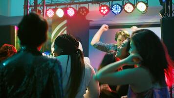 Crowd of people jumping on music, feeling happy on dance floor at nightclub. Young funky friends dancing on electronic music with DJ on stage, partying together with spotlights. photo