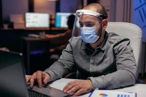 Businessman protect himself with visor against covid-19 late in the office distancing from colleagues. scrolling on laptop. Stressed man in corporation working hard to finish a project wearing face mask as safety precaution due to coronavirus pandemic. photo