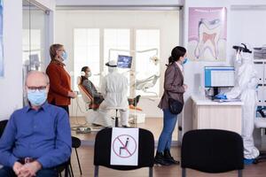 Patients with protection mask waiting in stomatology reception with new normal, staff wearing ppe suit. Woman checking appoiment with nurse coming for dental control during coronavirus pandemic photo