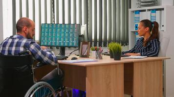 Businessman in wheelchair working in corporate office room changing documents analysing graphs. Employees in modern company typing on computer keyboard and pointing at desktop checking clients list. photo