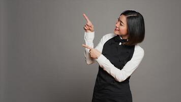 Asian waitress pointing up for an advertisement in studio, indicating direction above her head to create promotional ad. Woman catering employee points at something upwards. Camera A. photo