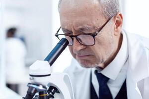 Close up of senior scientist analyzing virus sample using microscope in modern facility. Chemist researcher in sterile lab doing experiments for medical industry using modern technology. photo